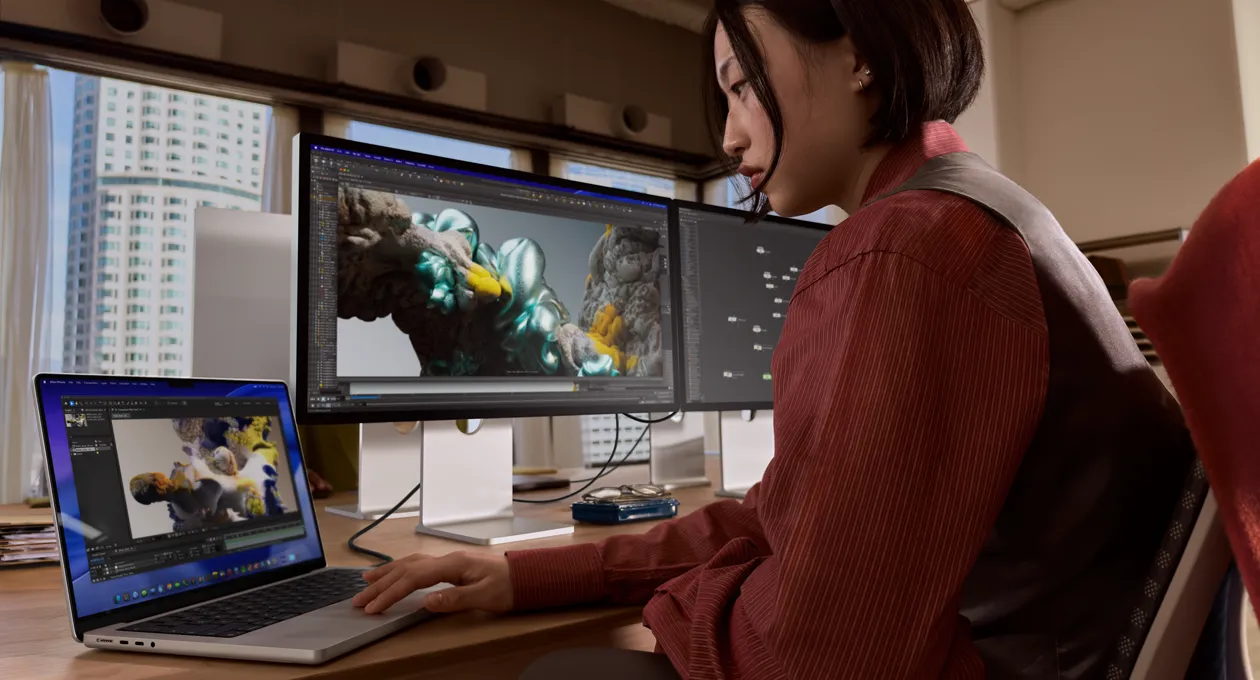 在办公室环境中，一个人在一台 MacBook 笔记本电脑上工作，笔记本电脑通过雷雳 5 端口串联 Studio Display XDR。